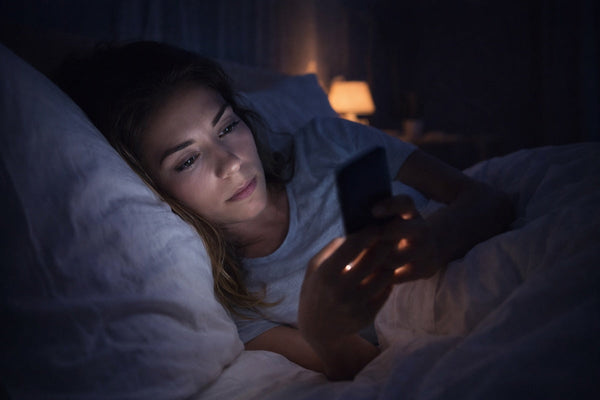 A person lying in bed at night illuminated by the bright light of a smartphone, illustrating how evening screen exposure can disrupt sleep and circadian rhythm.