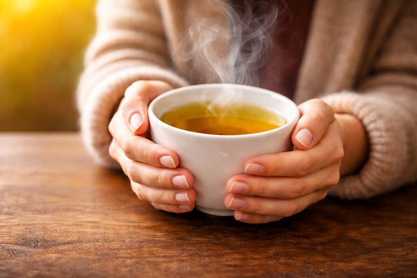 A close-up of a person’s hands holding a steaming cup of tea on a wooden table, symbolizing calm, focus, and the health benefits of tea.